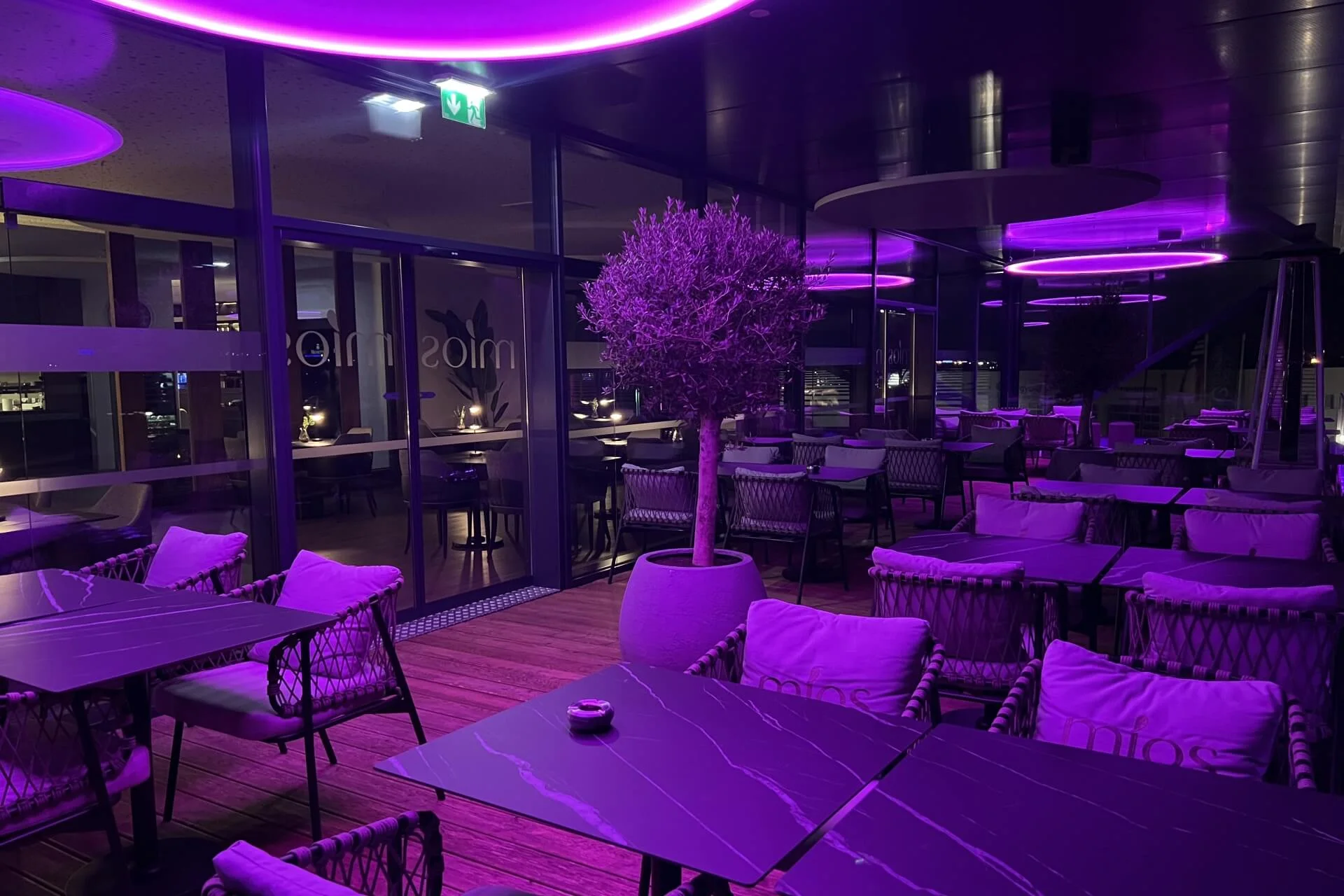 Modernes, überdachtes Restaurant mit leeren Tischen und Stühlen, beleuchtet von lila Neonröhren. In der Mitte steht ein getöpferter Baum, und große Glasfenster säumen die Rückwand und verleihen dem Raum eine stilvolle und moderne Atmosphäre.