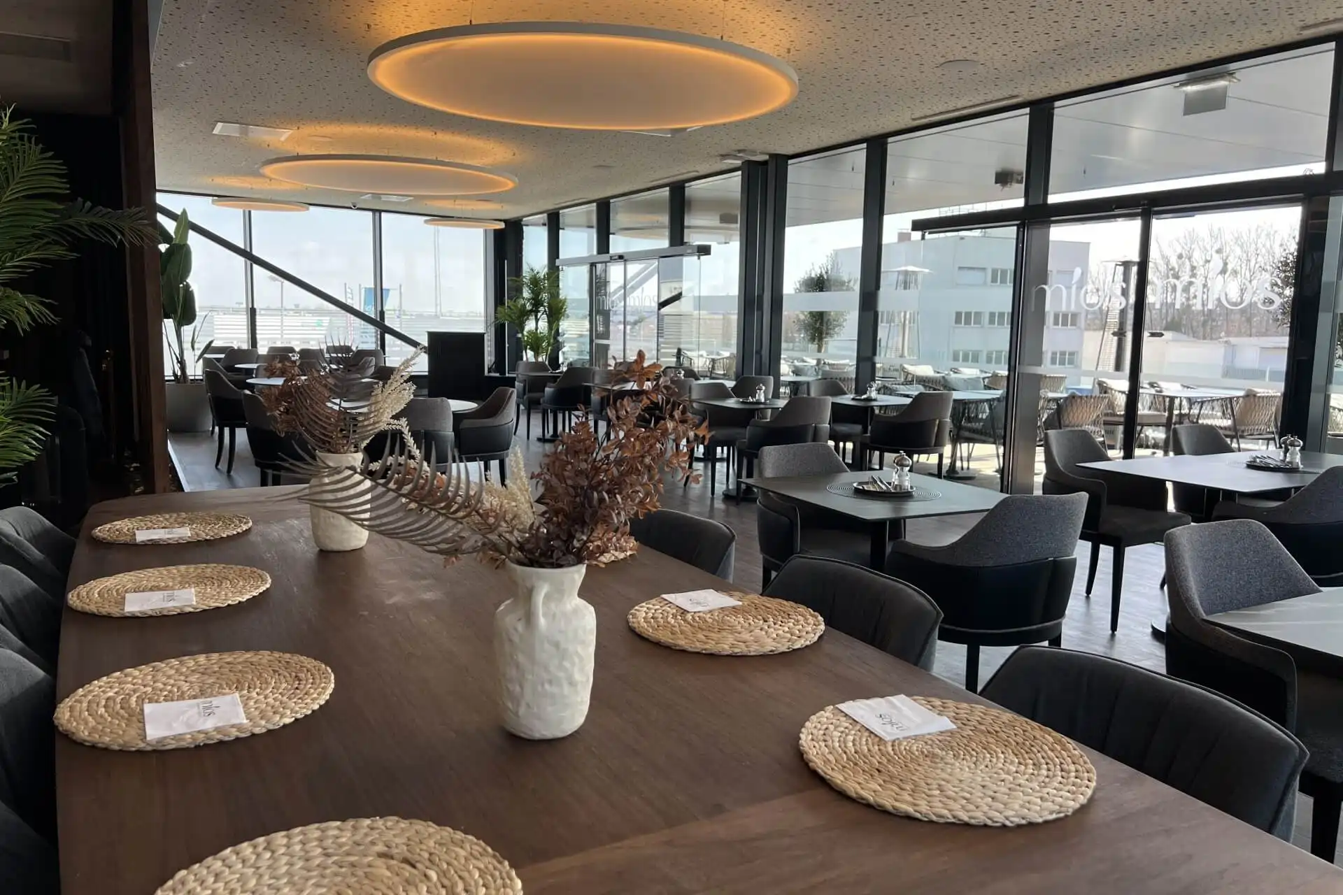 Modernes Restaurant mit großen Fenstern, Holztischen mit gewebten Tischsets, Vasen mit getrockneten Pflanzen und grauen gepolsterten Stühlen. Natürliches Licht erfüllt den Raum und bietet einen Blick auf die Gebäude der Stadt.