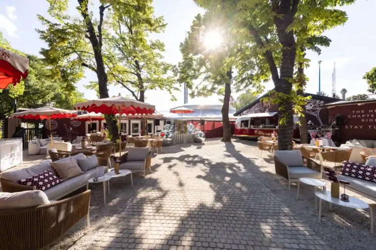 Mirage Garden Sonniges Straßencafé mit Korbstühlen, Tischen und rot-weißen Markisen. Bäume spenden Schatten, und das Sonnenlicht fällt durch das Laub auf eine Terrasse mit Kopfsteinpflaster. Entspannte, einladende Atmosphäre.