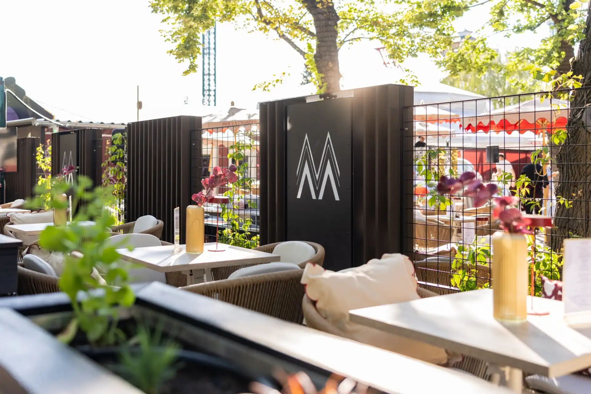 Terrasse eines Restaurants im Freien mit weißen Tischen und Stühlen, Topfpflanzen und einem schwarzen Zaun mit einem weißen Doppel-M-Logo. Das Sonnenlicht fällt durch die Bäume und schafft eine helle und einladende Atmosphäre.