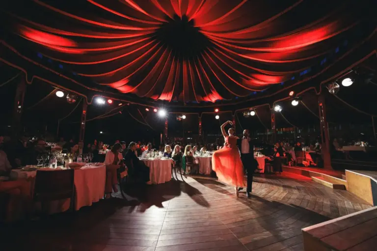 Ein Paar tanzt unter einer roten Zeltdecke bei einer formellen Veranstaltung. Die Gäste sitzen an runden Tischen mit weißen Tischtüchern und verfolgen die Aufführung in einem warm beleuchteten, eleganten Raum.