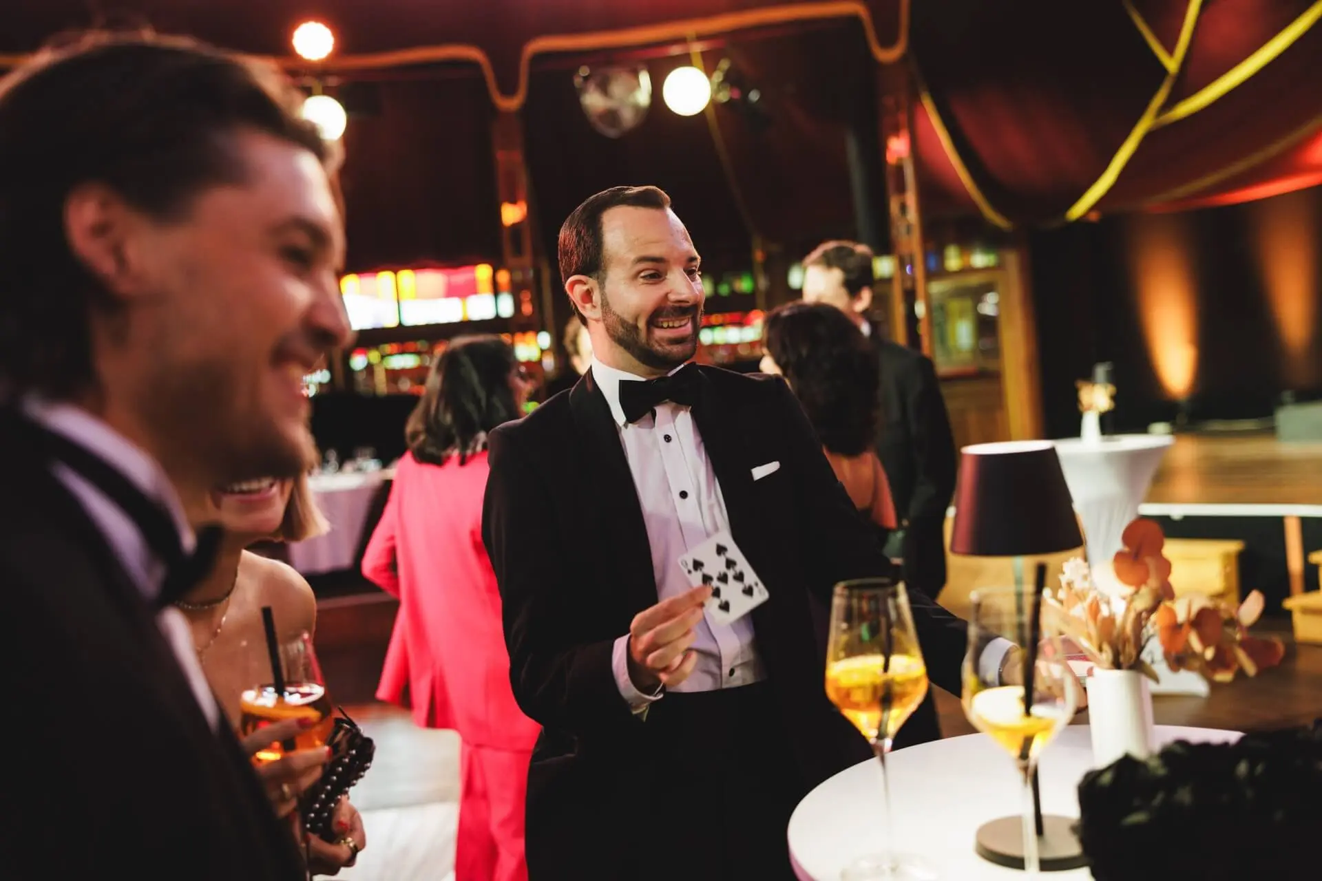 Ein Mann im Smoking hält Spielkarten in der Hand, während er lächelt und die Gäste bei einer formellen Veranstaltung unterhält. Die Menschen um ihn herum sind elegant gekleidet und halten Getränke in der Hand, was auf eine lebhafte und festliche Atmosphäre schließen lässt.