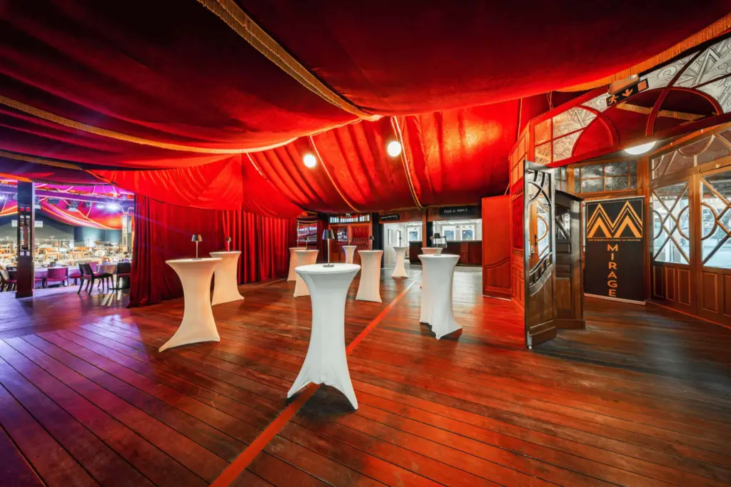 Luxuriöser Veranstaltungsraum mit reichlich rot drapierter Decke, Holzböden, Steh-Cocktailtischen mit weißen Hussen und einem offenen Eingang mit der Aufschrift Mirage. Im Hintergrund ist der Essbereich mit gedeckten Tischen zu sehen.