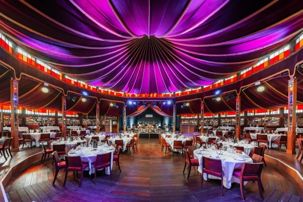 Ein großzügiger, kreisförmiger Speisesaal mit Holzfußboden, runden Tischen für die Gäste und einer dramatischen violetten Zeltdecke, die durch rote, bernsteinfarbene und blaue Glasmalereien betont wird. Am hinteren Ende ist eine Bühne zu sehen.