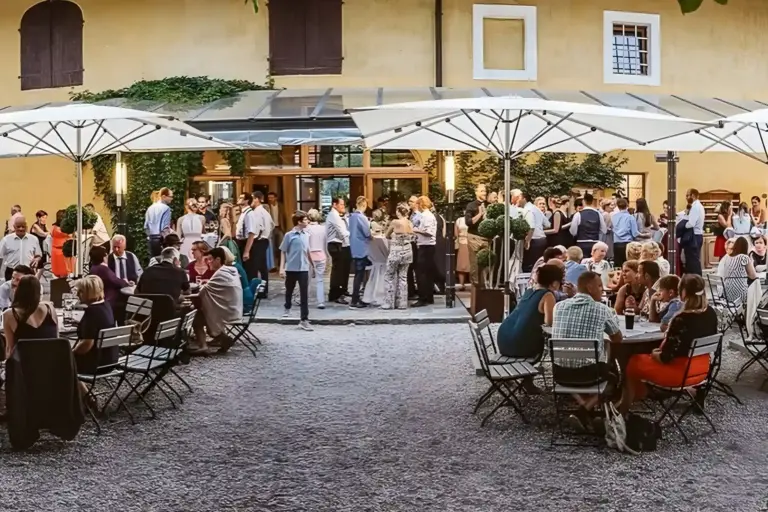 Versammlung im Freien in einem Hofrestaurant, wo die Leute an Tischen sitzen und andere stehen und sich unter großen weißen Sonnenschirmen mischen, umgeben von einem gelben Gebäude mit Grünpflanzen.