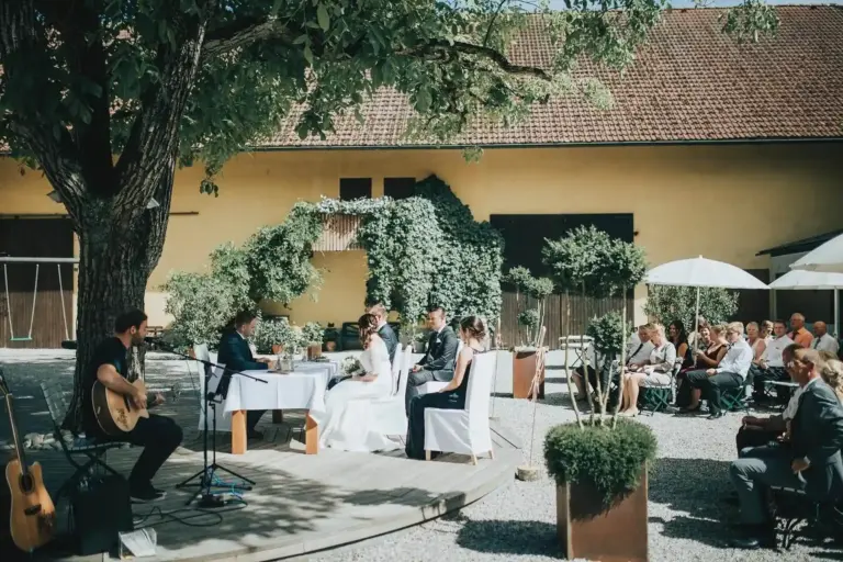 Die Trauung findet im Freien unter einem großen Baum statt, wobei Braut und Bräutigam an einem Tisch Platz nehmen. Die Gäste sitzen in der Nähe, und ein Musiker mit einer Gitarre spielt auf einer kleinen Holzbühne. Im Hintergrund ist ein gelbes Gebäude zu sehen.