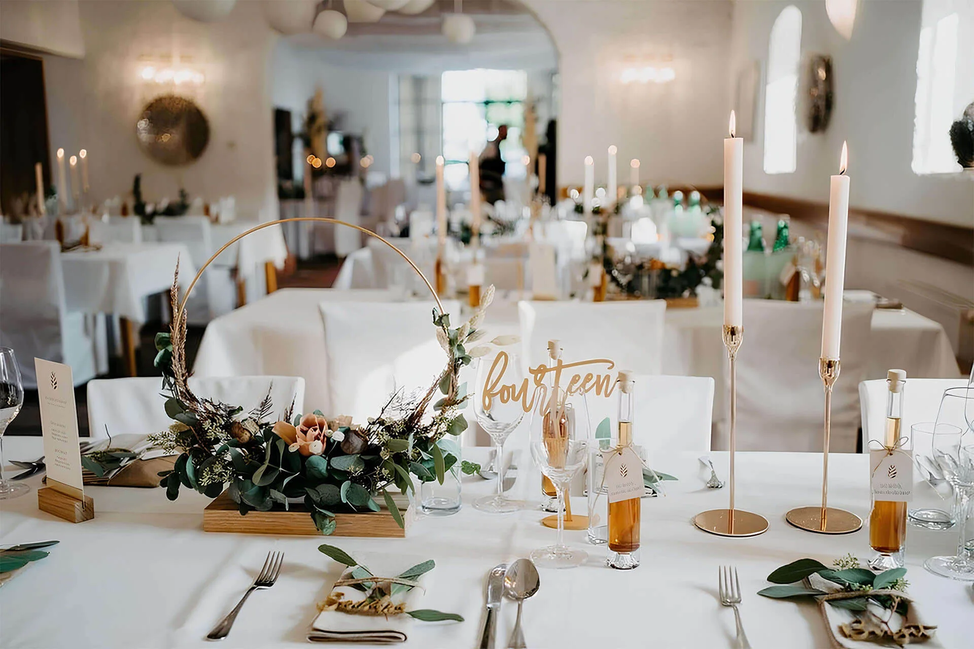 Eine Hochzeitstafel ist mit eleganter weißer Bettwäsche, goldenen Kerzenhaltern mit hohen Kerzen, grünen Tafelaufsätzen, kleinen Flaschen als Gastgeschenke und einem Tischnummernschild mit der Aufschrift "vierzehn" geschmückt. Im Hintergrund sind weitere dekorierte Tische zu sehen.