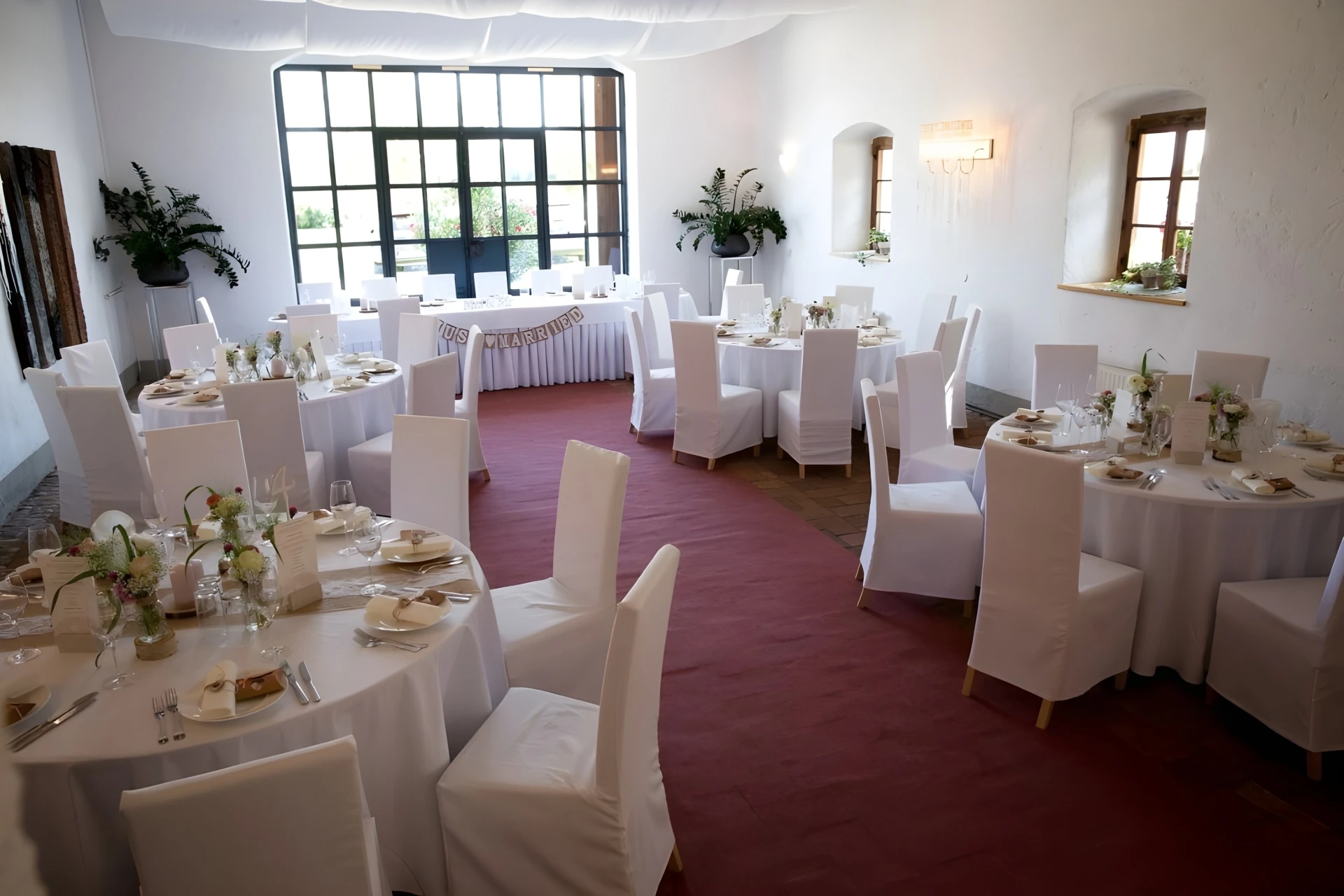 Ein heller, eleganter Bankettsaal mit runden, weiß gedeckten Tischen, schlichten Blumenkränzen und weißen Stuhlhussen. Große Fenster lassen natürliches Licht herein, und der Raum ist für eine formelle Veranstaltung oder einen Hochzeitsempfang eingerichtet.