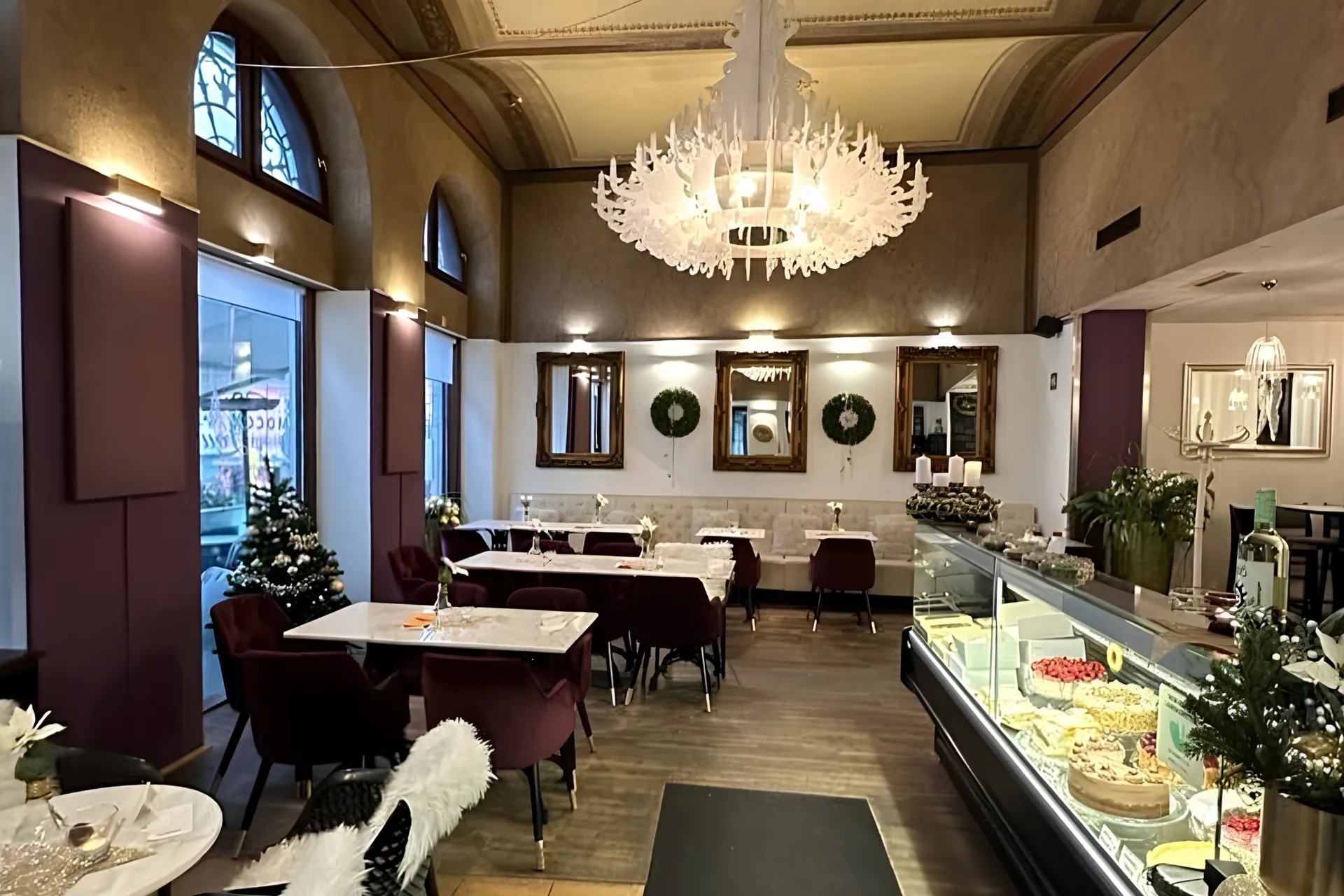 Ein elegantes Restaurantinterieur mit einem großen weißen Kronleuchter, Tischreihen mit weichen violetten Stühlen und einer Glasvitrine voller Kuchen.