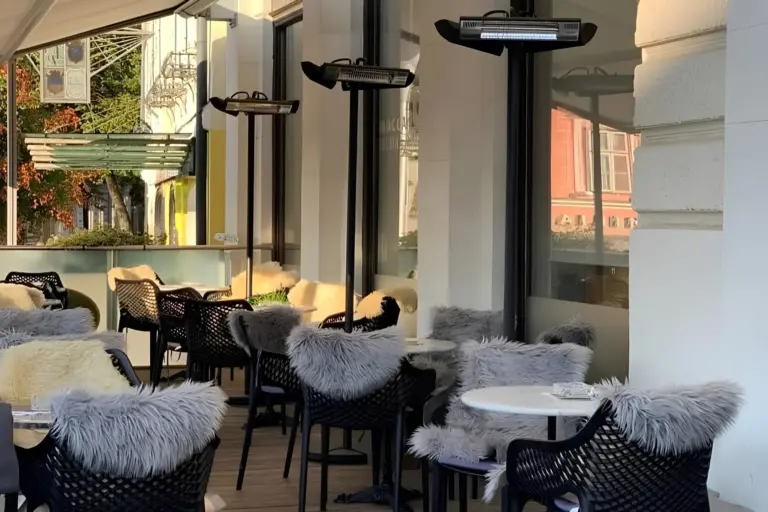 Café-Sitzbereich im Freien mit schwarzen Stühlen, die mit grauen und weißen Kunstfellkissen bezogen sind, kleinen runden Tischen und Terrassenheizstrahlern.