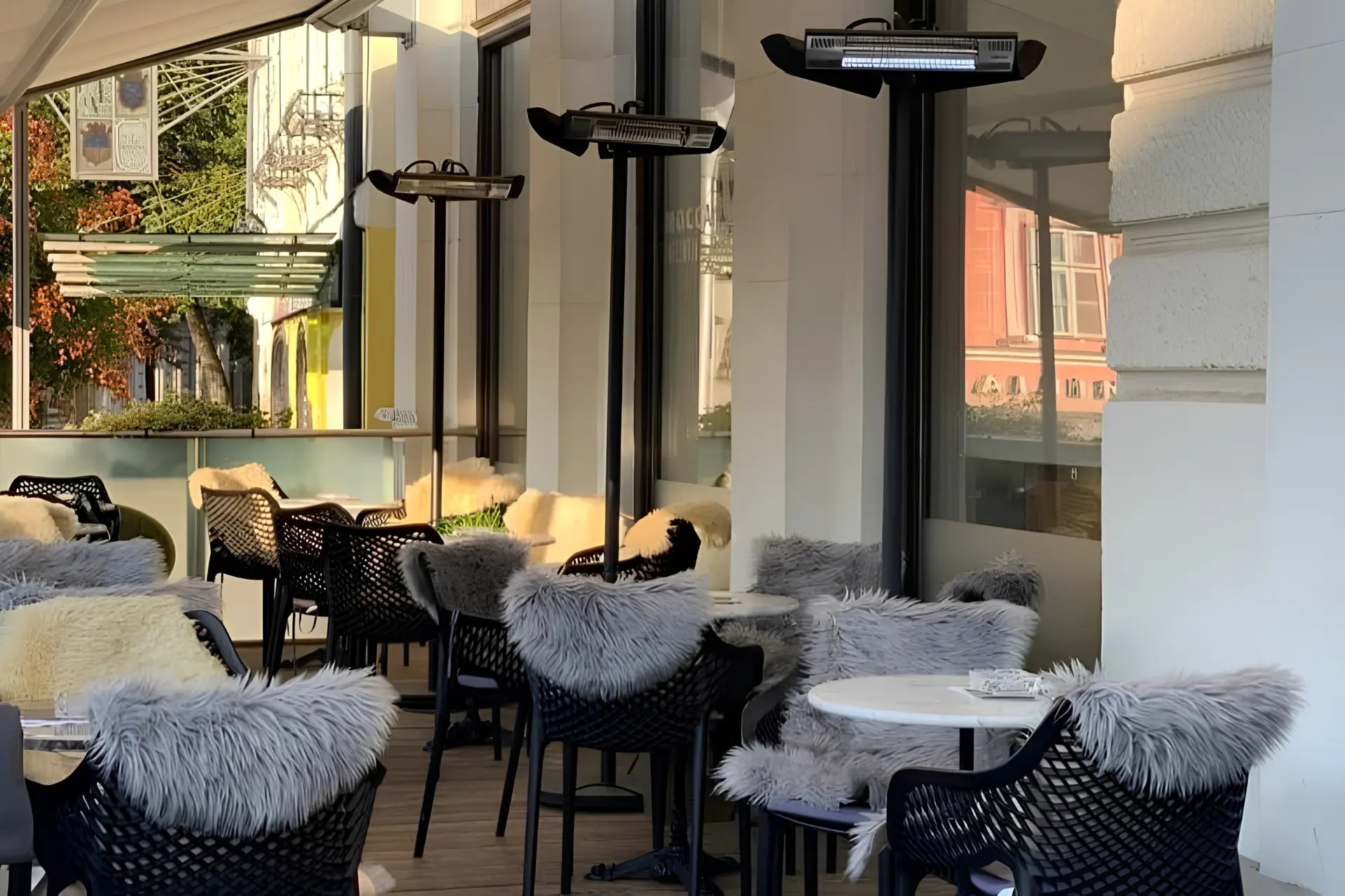 Café-Sitzbereich im Freien mit schwarzen Stühlen, die mit grauen und weißen Kunstfellkissen bezogen sind, kleinen runden Tischen und Terrassenheizstrahlern.