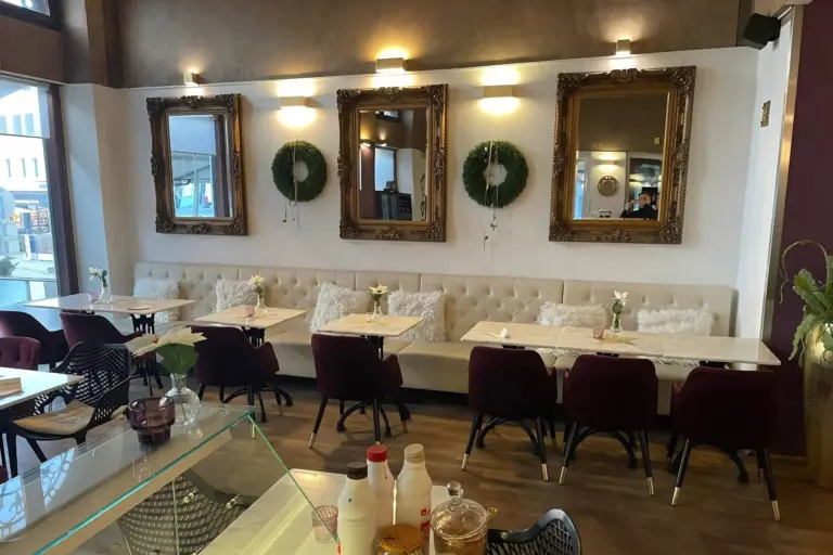 Das Interieur eines Cafés mit einer langen, cremefarbenen, gepolsterten Sitzecke mit passenden Stühlen, Tischen mit Marmorplatten und drei goldgerahmten Spiegeln, die zwischen zwei grünen Kränzen an der Wand hängen.