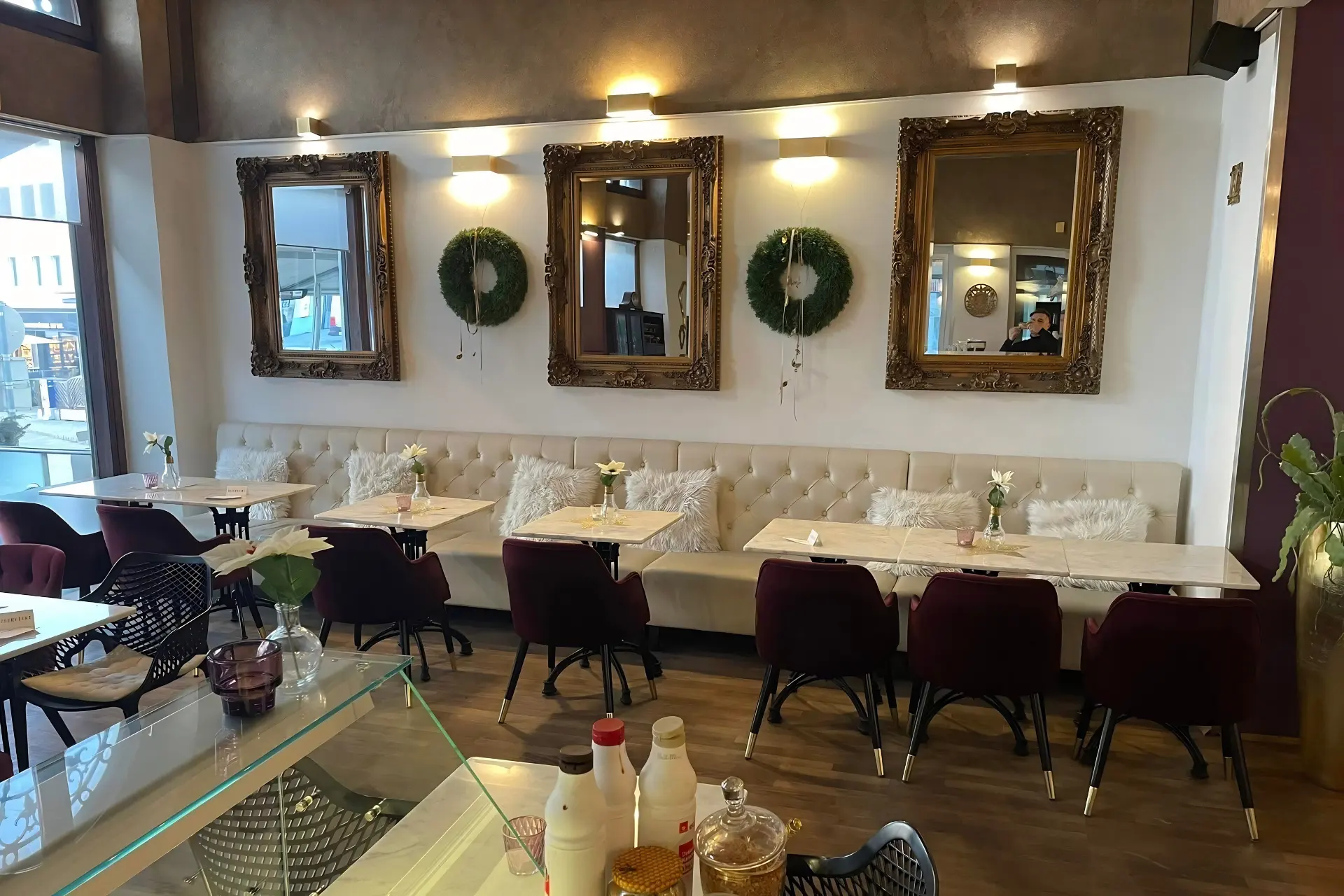 Das Interieur eines Cafés mit einer langen, cremefarbenen, gepolsterten Sitzecke mit passenden Stühlen, Tischen mit Marmorplatten und drei goldgerahmten Spiegeln, die zwischen zwei grünen Kränzen an der Wand hängen.