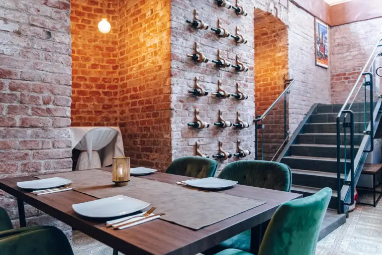 Ein Restaurant mit freiliegenden Backsteinwänden, einem mit Tellern und Besteck gedeckten Tisch, grünen Samtstühlen, einer Kerze in der Mitte, Weinflaschen an der Wand und einer Treppe, die in eine obere Etage führt.