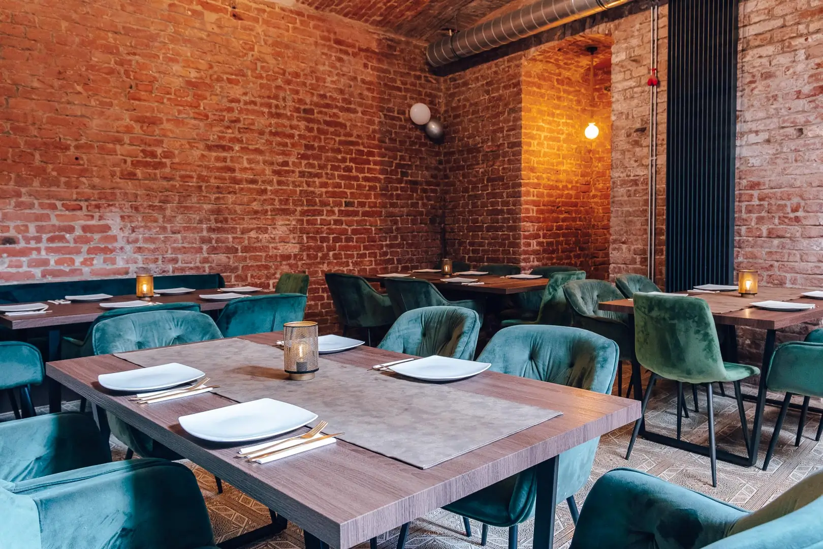 Ein gemütliches Restaurant mit freiliegenden Backsteinwänden, Holztischen mit weißen Tellern und Besteck, grünen Samtstühlen und warmer Beleuchtung.