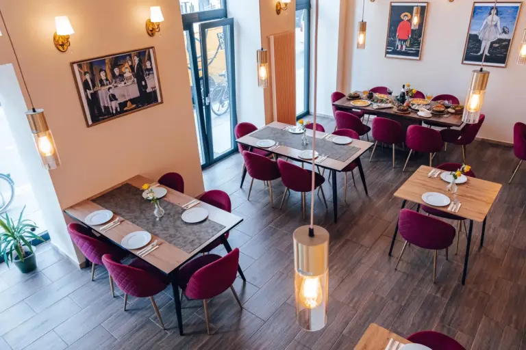 Ein modernes Restaurant mit roten Samtstühlen, gedeckten Tischen mit weißen Tellern und Gläsern, Holzböden, Wandkunst, großen Fenstern und Hängelampen.