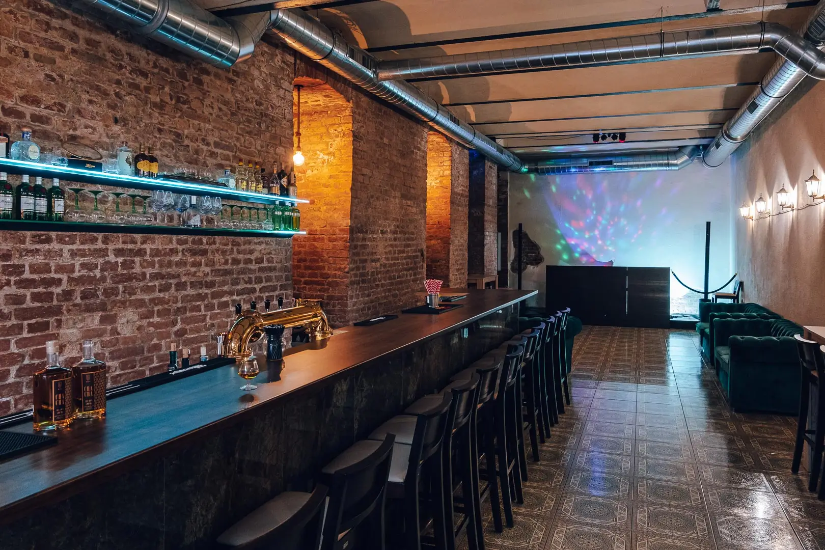 Eine moderne Bar mit freiliegenden Backsteinwänden, einem langen Holztresen mit Barhockern, Regalen mit Flaschen, warmer Beleuchtung und einer kleinen Bühne mit bunten Lichtern am anderen Ende.