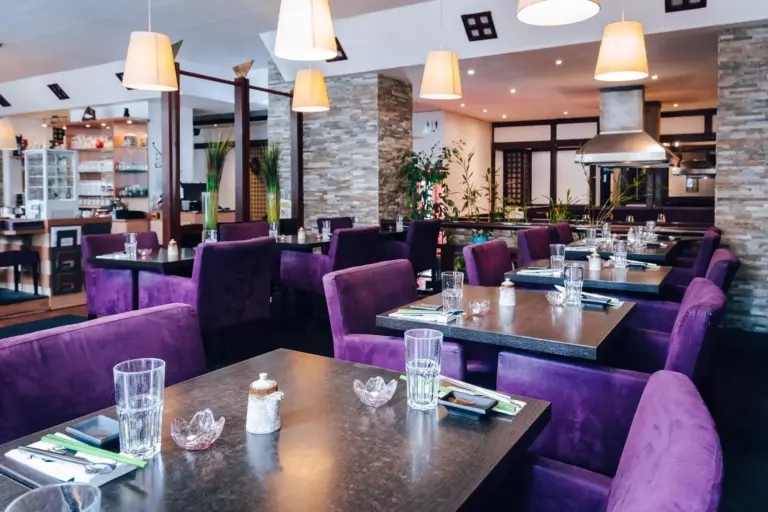 Ein moderner Restaurantbereich mit lila gepolsterten Stühlen, ordentlich gedeckten Tischen mit Gläsern, Besteck und Servietten und warmen Hängelampen, die eine gemütliche Atmosphäre schaffen. Pflanzen und dekorative Wände ergänzen die stilvolle Einrichtung.