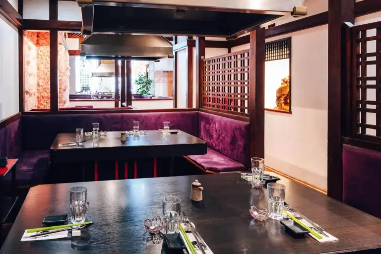 Ein modernes japanisches Restaurant mit dunklen Holztischen, die mit Stäbchen, Gläsern und Servietten eingedeckt sind, umgeben von lilafarbenen gepolsterten Sitzmöbeln und Holzgitterdekor unter einer Grillhaube aus Edelstahl.