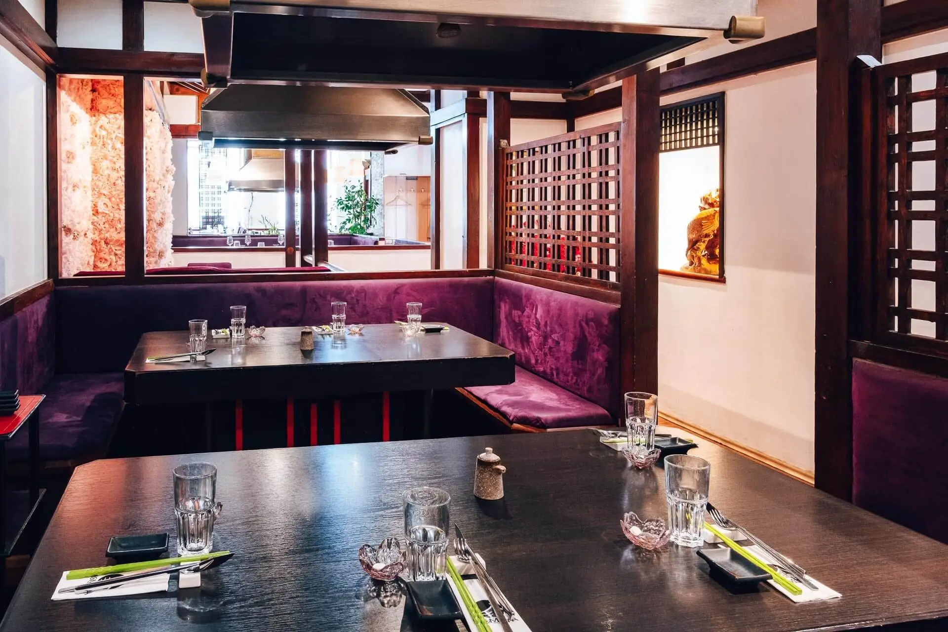 Ein modernes japanisches Restaurant mit dunklen Holztischen, die mit Stäbchen, Gläsern und Servietten eingedeckt sind, umgeben von lilafarbenen gepolsterten Sitzmöbeln und Holzgitterdekor unter einer Grillhaube aus Edelstahl.