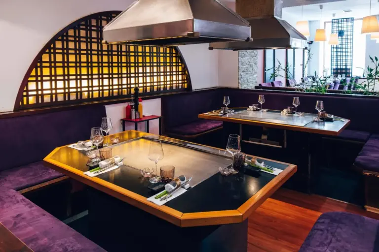 Ein modernes Restaurant mit zwei Teppanyaki-Tischen, lilafarbenen, gepolsterten Sitzgelegenheiten, Tischen mit Gläsern und Besteck sowie sanfter Beleuchtung durch Deckenlampen und einen dekorativen, hinterleuchteten Holzschirm.