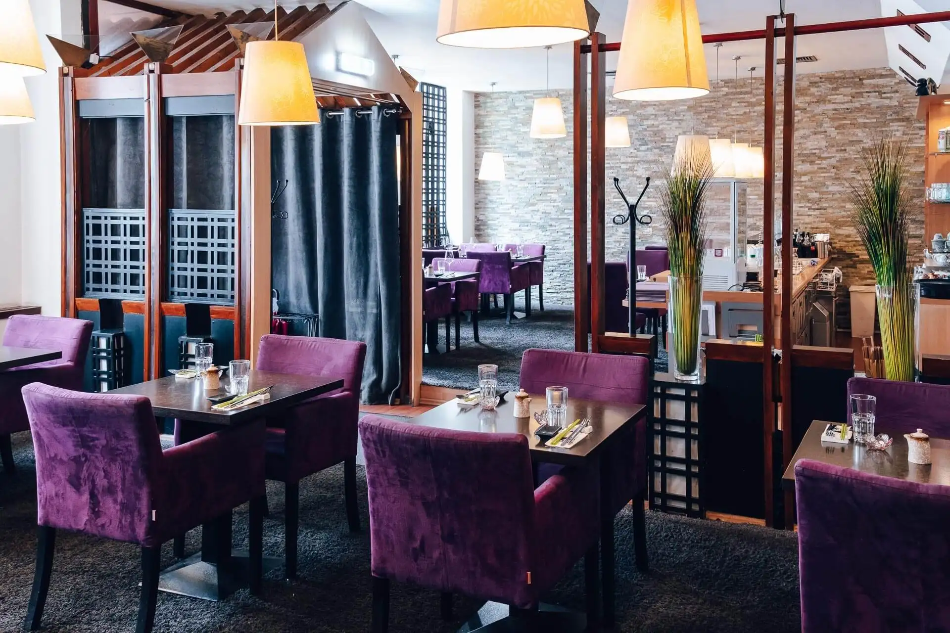 Ein modernes Restaurant mit violetten Samtstühlen, dunklen Holztischen mit Gläsern und Geschirr, zartgelben Hängelampen und abgetrennten Essbereichen mit dekorativen Paravents und hohen Vasen.