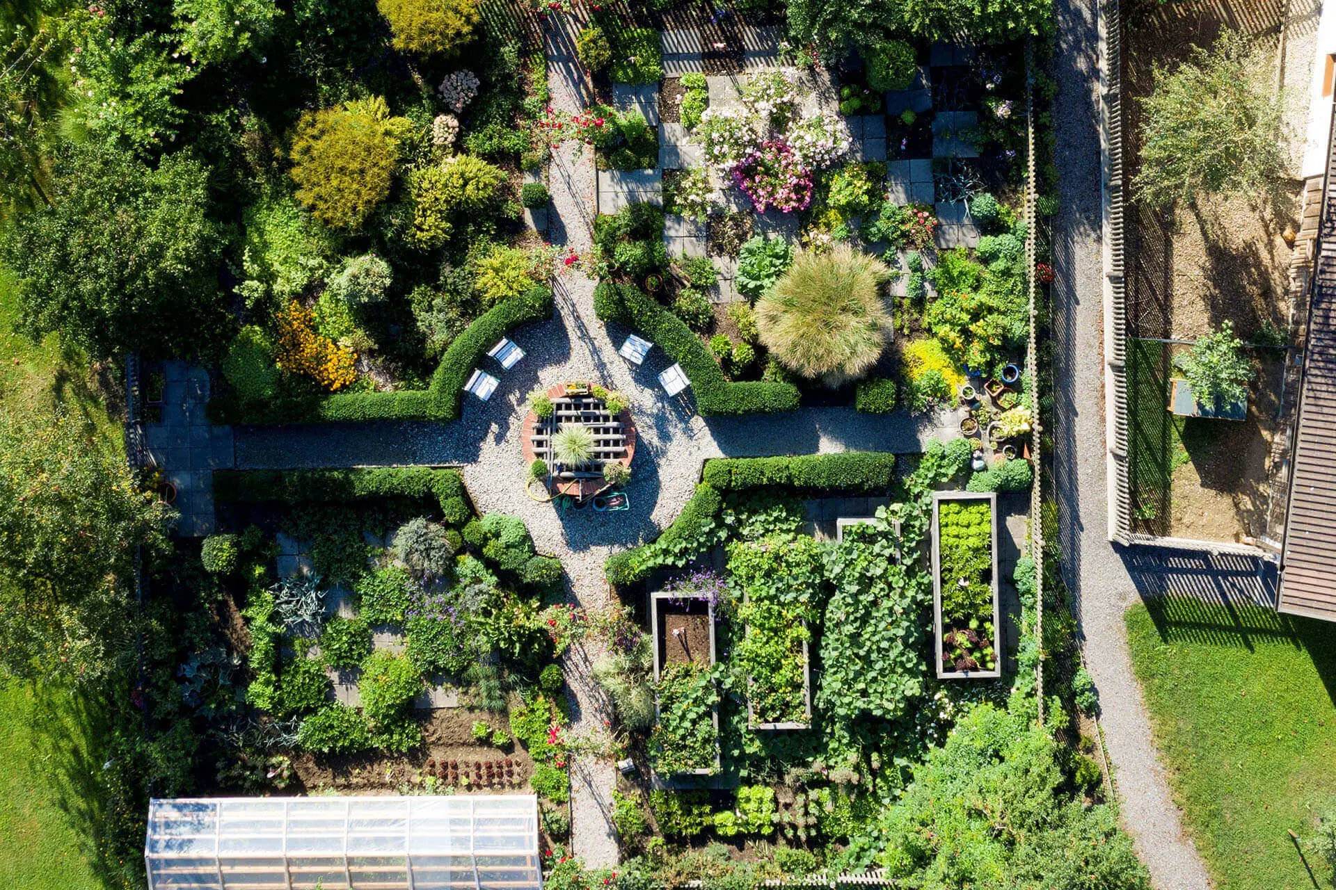 Luftaufnahme eines gepflegten Gartens mit geometrischen Wegen, Hochbeeten, üppigem Grün, Blumenbeeten, einem runden Sitzbereich in der Mitte und einem Gewächshaus in der unteren linken Ecke.