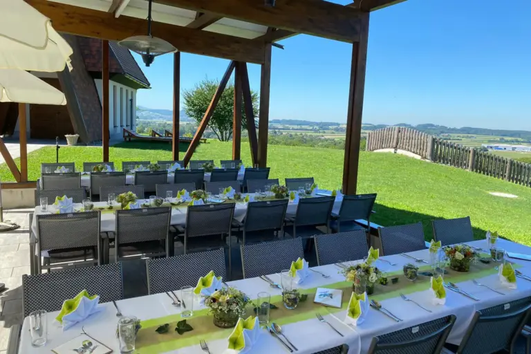 Essbereich im Freien mit langen Tischen für eine Mahlzeit, dekoriert mit grünen Akzenten und Blumenarrangements, unter einer Pergola aus Holz. Von hier aus hat man einen Blick auf den Rasen und die malerische Landschaft unter dem blauen Himmel.
