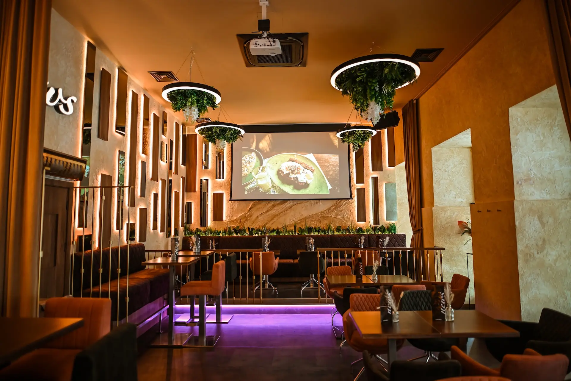 News Modernes Restaurantinterieur mit warmer Beleuchtung, von der Decke hängenden Pflanzen, gepolsterten Stühlen, Tischen und einem großen Wandbildschirm, auf dem die Speisen angezeigt werden. Die Einrichtung umfasst vertikale Lichtpaneele und einen lila beleuchteten Bodenbereich.