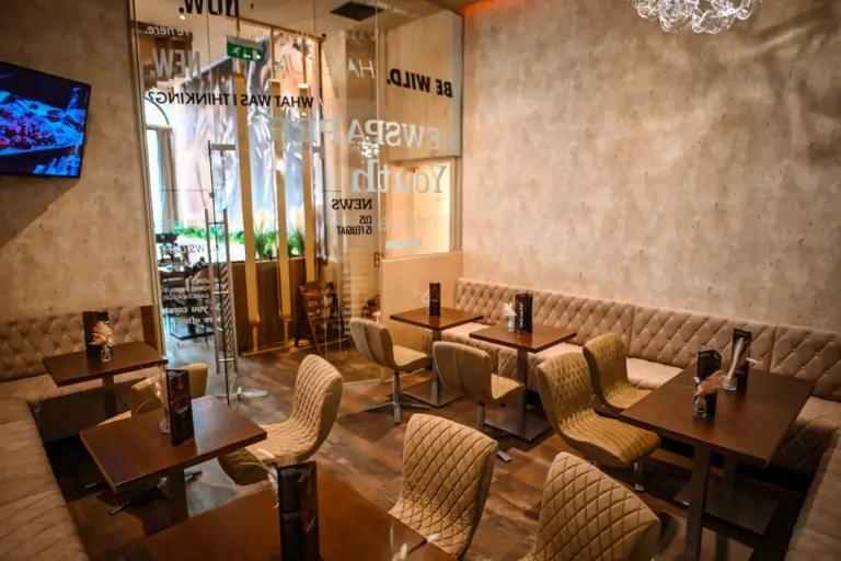 Ein gemütliches Café mit hellbraunen, gesteppten Stühlen, Holztischen, beigen, gepolsterten Bänken und dekorativen Wandtexten. Natürliches Licht fällt durch große Glastüren ein und schafft eine warme Atmosphäre.