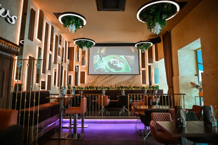 Ein modernes Restaurant mit stilvoller Beleuchtung, hängenden Pflanzen, gepolsterten Sitzgelegenheiten und einem großen Bildschirm, auf dem die Speisen angezeigt werden. Der Raum bietet eine Mischung aus Tischen und Ständen, warme Farbtöne und eine moderne Einrichtung.