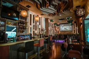 Modernes Barinterieur mit stilvoller Beleuchtung, hohen Stühlen um eine lange Bar, hängenden Pflanzen, dekorativen Deckenpaneelen und einem großen Bildschirm, auf dem die Speisen angezeigt werden. Die warme Beleuchtung schafft eine gemütliche, gehobene Atmosphäre.