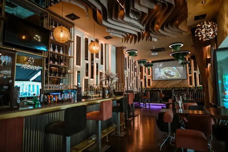 Modernes Barinterieur mit stilvoller Beleuchtung, hohen Stühlen um eine lange Bar, hängenden Pflanzen, dekorativen Deckenpaneelen und einem großen Bildschirm, auf dem die Speisen angezeigt werden. Die warme Beleuchtung schafft eine gemütliche, gehobene Atmosphäre.