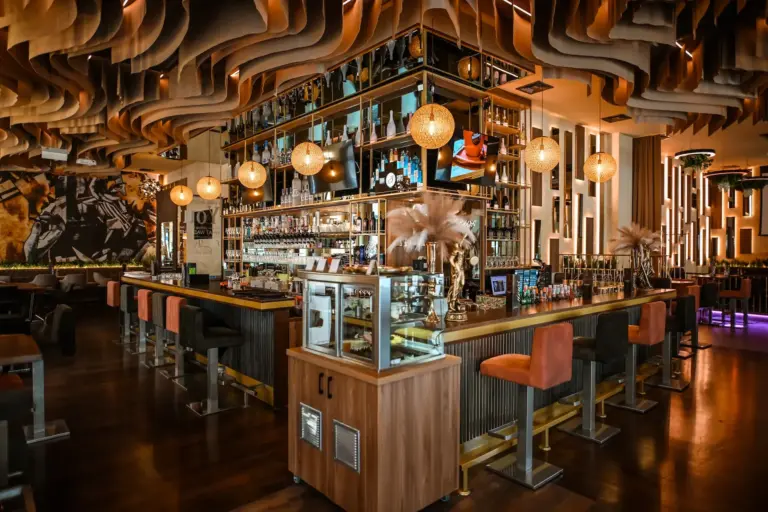 Eine stilvolle Bar mit modernem Dekor, einer zentralen Inselbar, Regalen mit Flaschen, warmer Hängebeleuchtung, Barhockern aus Samt und gewellten Holzdecken, die eine gemütliche, gehobene Atmosphäre schaffen.