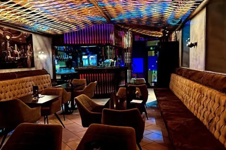 Eine moderne Lounge-Bar mit Plüschsesseln und -sofas, gedämpfter Beleuchtung und einer bunten, gemusterten Decke. Der Barbereich ist mit blauen und violetten Lichtern beleuchtet, die eine gemütliche und doch lebendige Atmosphäre schaffen.