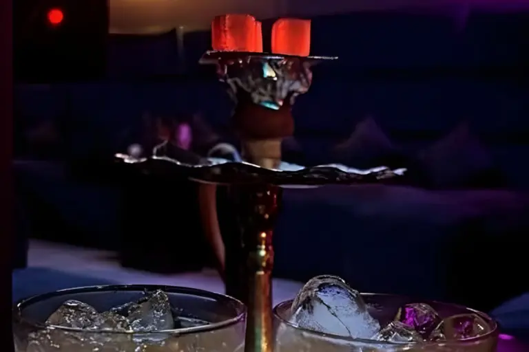 Eine Wasserpfeife mit glühenden Kohlen, die zwischen zwei mit Eiswürfeln gefüllten Gläsern steht. Der Hintergrund ist mit dunkelblauen und violetten Tönen schummrig beleuchtet und schafft eine entspannte Lounge-Atmosphäre.