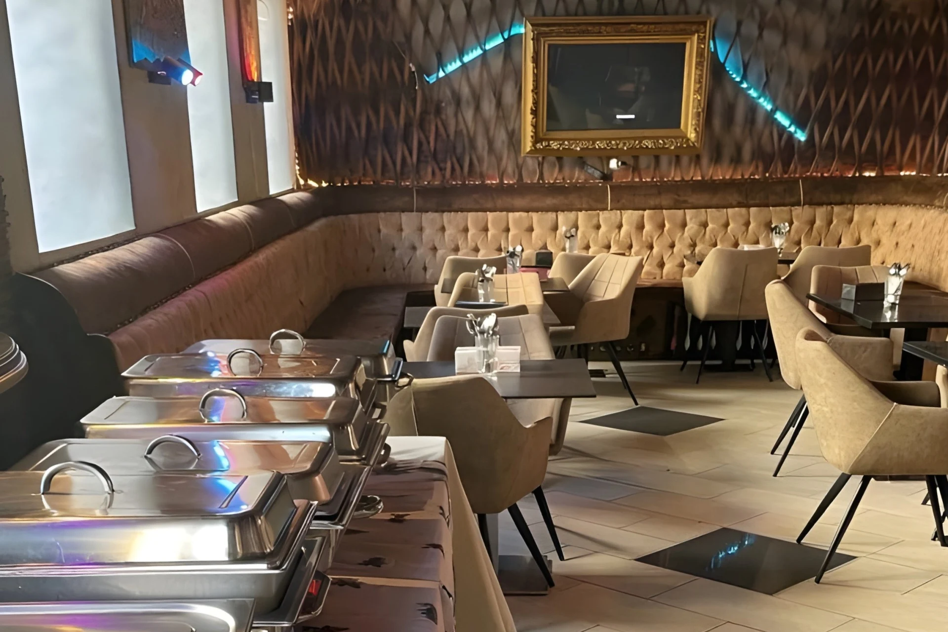 Ein modernes Restaurant mit beigefarbenen, getufteten Sitzgelegenheiten, leeren beigen Stühlen, Tischen mit Servietten und Gewürzen, Buffetschalen im Vordergrund und einem gerahmten Spiegel an einer gemusterten Wand.