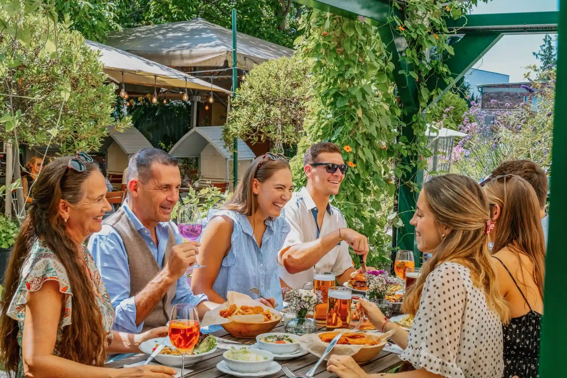 Eine Gruppe von sechs Personen sitzt um einen Tisch im Freien, lächelt und genießt eine gemeinsame Mahlzeit. Der Tisch ist mit Speisen und Getränken gefüllt, und die Umgebung ist üppig und grün mit Pflanzen und Bäumen im Hintergrund.