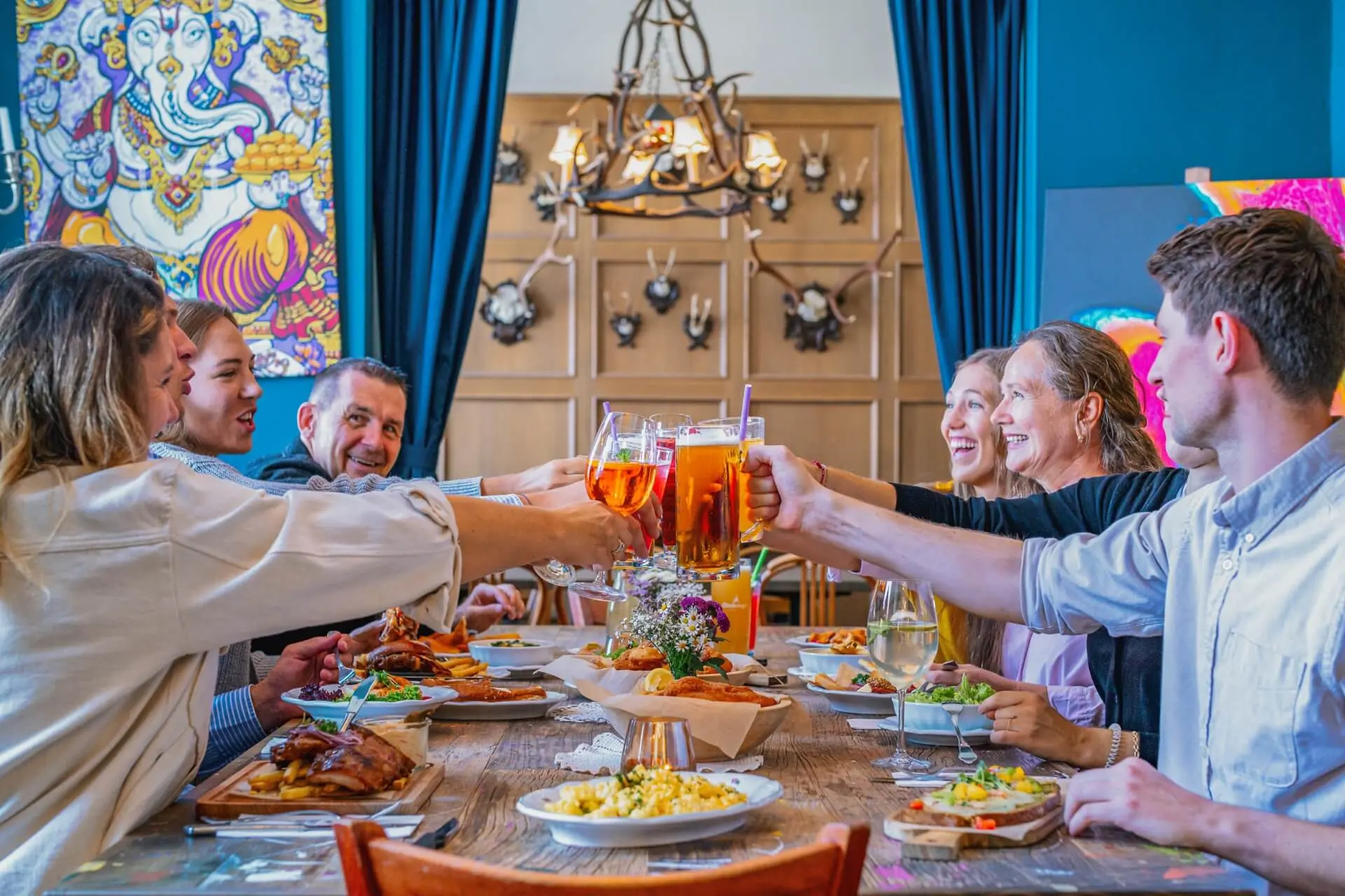 Eine Gruppe von sechs Personen sitzt um einen Tisch herum, lächelt und stößt bei einer Mahlzeit mit Getränken an. Der Tisch ist mit Essen gefüllt, und der Raum ist mit farbenfroher Kunst und Wanddekoration geschmückt.