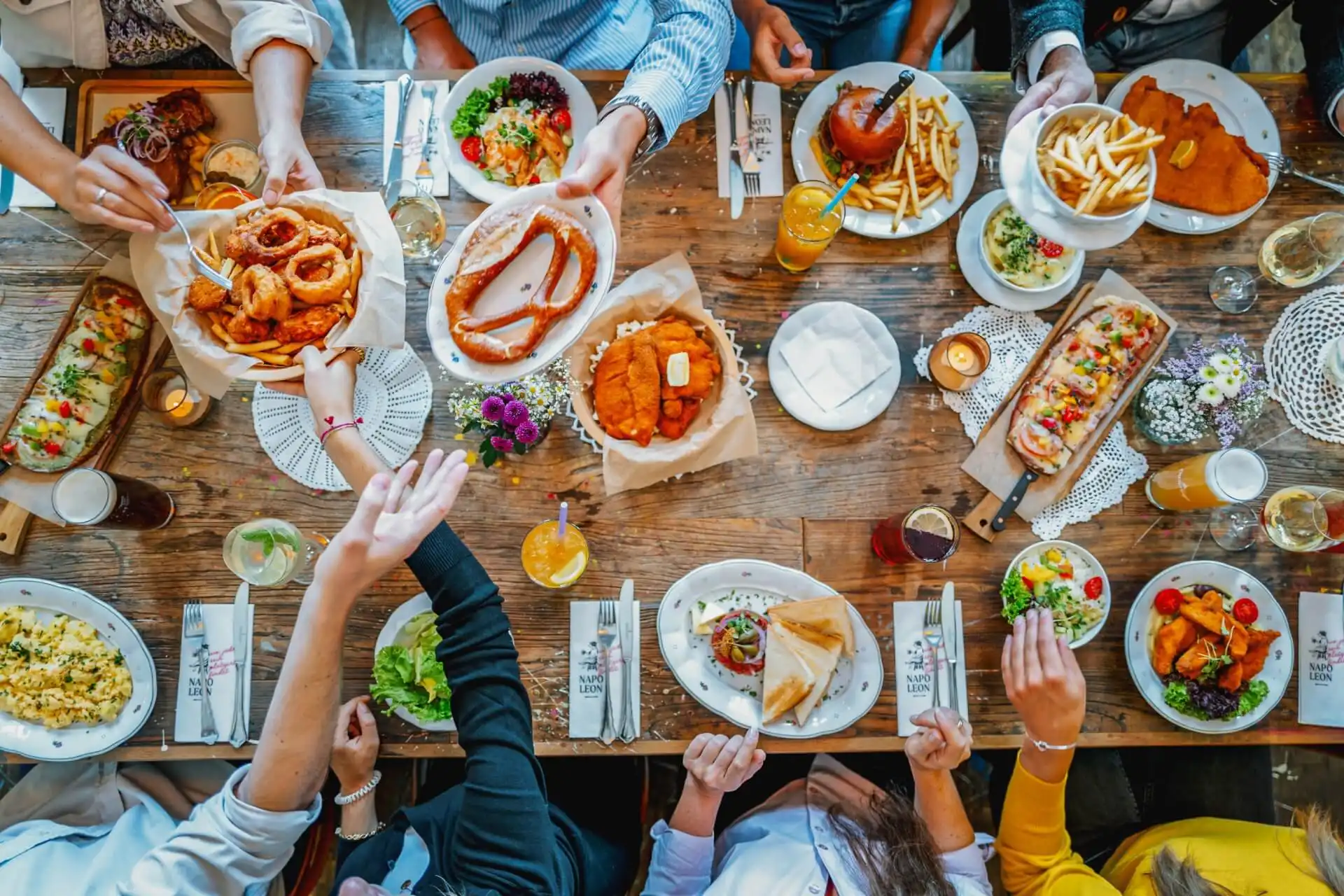 Die Menschen versammelten sich um einen Holztisch mit verschiedenen Gerichten, darunter Pommes frites, Salate, Schnitzel, Würstchen und Getränke, griffen nach dem Essen und teilten eine bunte, gemeinsame Mahlzeit in einer lebhaften Umgebung.