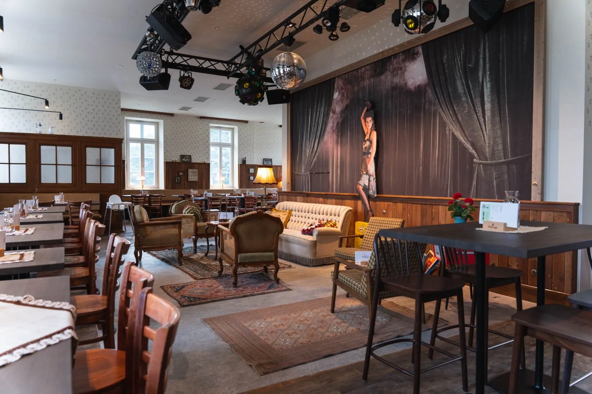 Ein gemütliches, stilvolles Café mit Holztischen, alten Stühlen, Teppichen und einer Bühne mit einem großen Wandgemälde einer Frau im Kostüm. Die warme Beleuchtung und das eklektische Dekor schaffen eine einladende Atmosphäre.