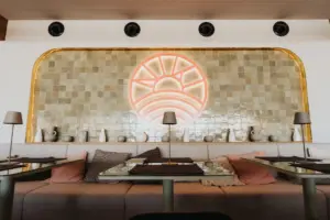 Modernes Restaurantinterieur mit gepolsterten Sitzbänken, dekorativen Kissen, minimalistischen Tischlampen und einer gekachelten Wand mit einem großen leuchtenden Neonsonnensymbol. Neutrale Farben und schlichte Töpferwaren schaffen eine gemütliche, stilvolle Atmosphäre.