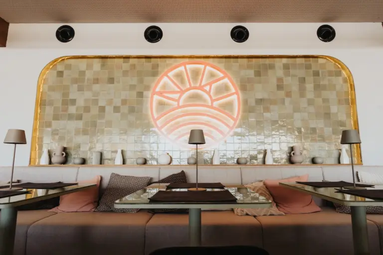 Modernes Restaurantinterieur mit gepolsterten Sitzbänken, dekorativen Kissen, kleinen Tischlampen und einer gekachelten Wand mit einem großen Neon-Sonnenaufgangsmotiv. Vasen und Dekorationsartikel sind entlang eines Regals unterhalb der Neonkunst angeordnet.