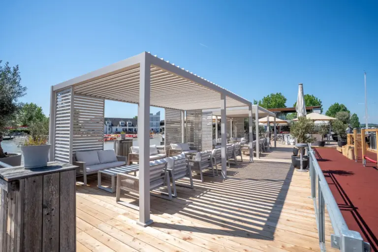 Moderner Freisitz mit weißen Pergolen, gepolsterten Stühlen und Tischen auf einer Holzterrasse unter strahlend blauem Himmel; im Hintergrund sind Wasser und Häuser zu sehen.