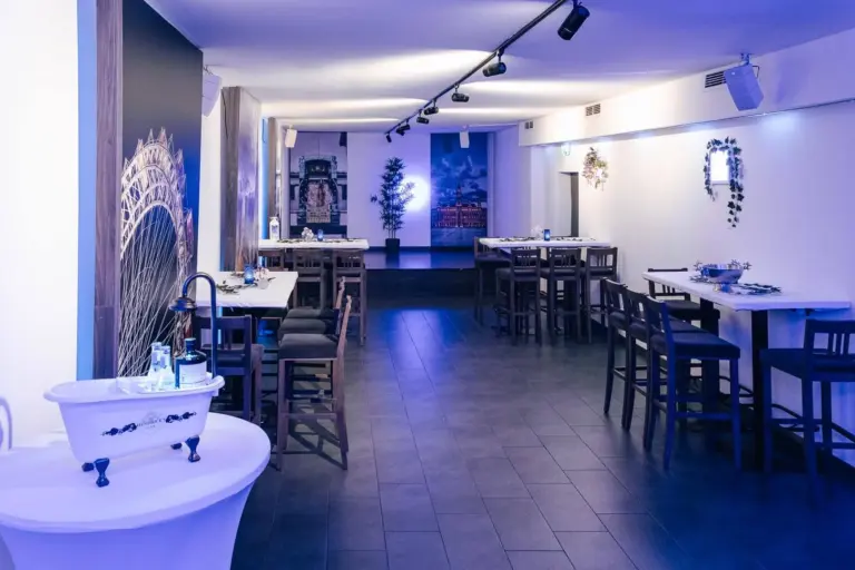 Ein modernes, schummriges Restaurant mit hohen Tischen und Stühlen, minimalistischem Dekor und Wandkunst. Die Tische sind auf beiden Seiten angeordnet, mit einem weißen, wannenförmigen Waschbecken im Vordergrund und Pflanzen und Kunst an den Wänden.