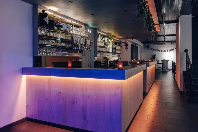 Eine moderne Bar mit einem hölzernen Tresen, der von sanften violetten und orangefarbenen Lichtern beleuchtet wird, darüber Regale mit Gläsern und Flaschen und im Hintergrund Happy-Birthday-Banner. Tische und Stühle säumen den gefliesten Boden.