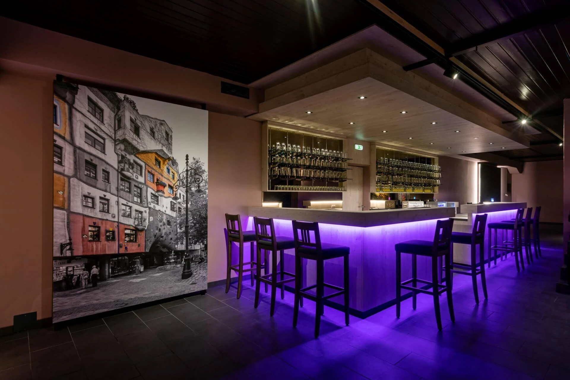 Eine moderne Bar mit lilafarbener Unterbeleuchtung, hohen Stühlen um die Bar herum, Flaschenregalen dahinter und einem großen schwarz-weißen Wandgemälde mit einem Hauch von Farbe an der linken Wand. Der Raum ist spärlich beleuchtet und stilvoll.