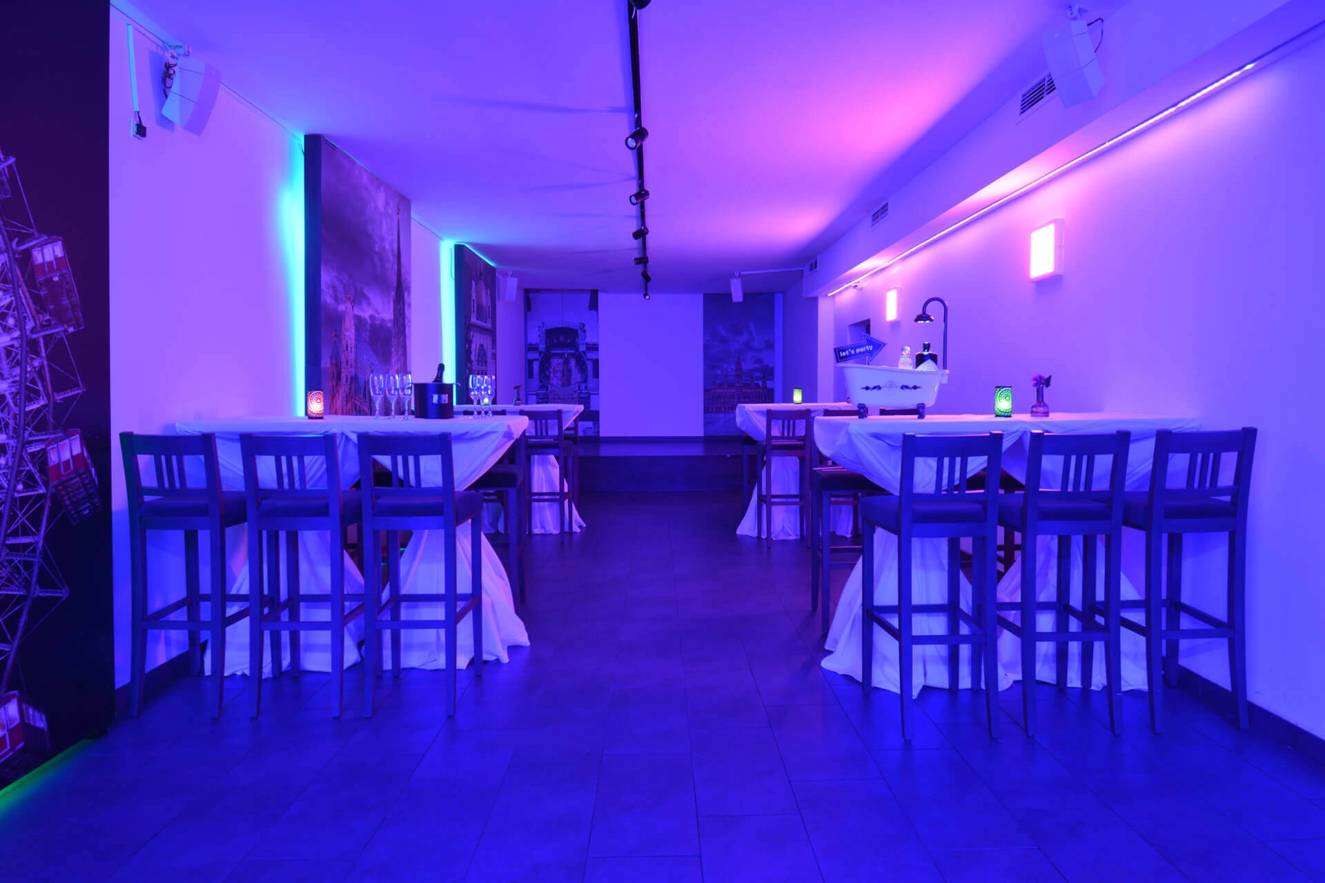 Eine moderne Bar oder Lounge mit hohen Tischen und Stühlen, beleuchtet von blauer und violetter Umgebungsbeleuchtung. Die Tische sind mit Tischtüchern bedeckt, und Kerzen oder Lichter sorgen für eine gemütliche Atmosphäre in dem stilvollen, modernen Raum.