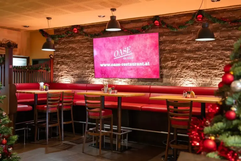 Oase am Teich Ein gemütliches Restaurant mit roten Polstersesseln, Holztischen, Weihnachtsdekoration und einem großen Bildschirm, auf dem „OASE“ und eine Website-URL an einer Wand mit Steinakzenten zu sehen sind.