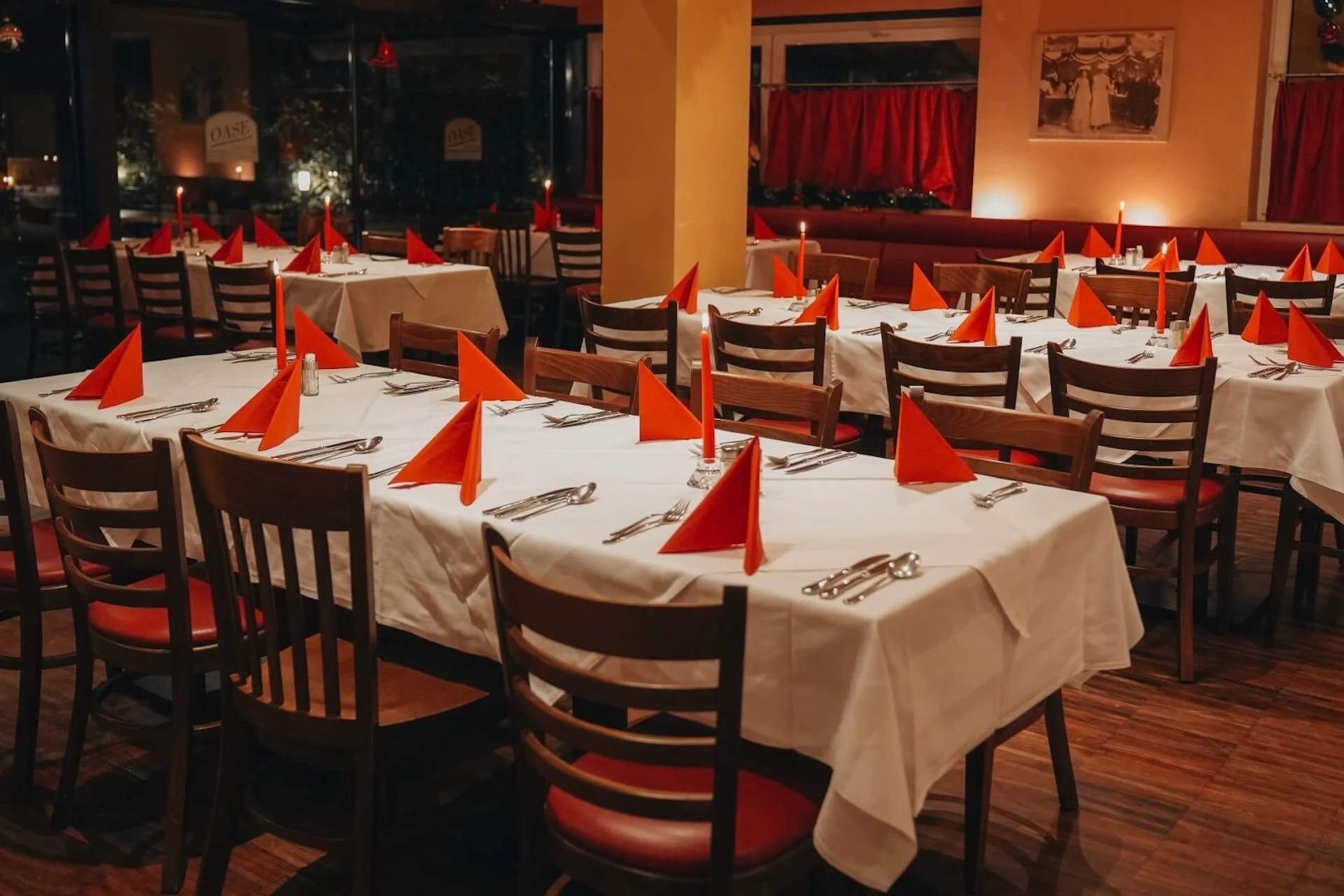 Ein leeres Restaurant mit Holzstühlen und Tischen, die mit weißen Tischdecken gedeckt sind; auf jedem liegen rot gefaltete Servietten, Teller und Besteck. Warme Beleuchtung und rote Vorhänge sorgen für eine gemütliche Atmosphäre.